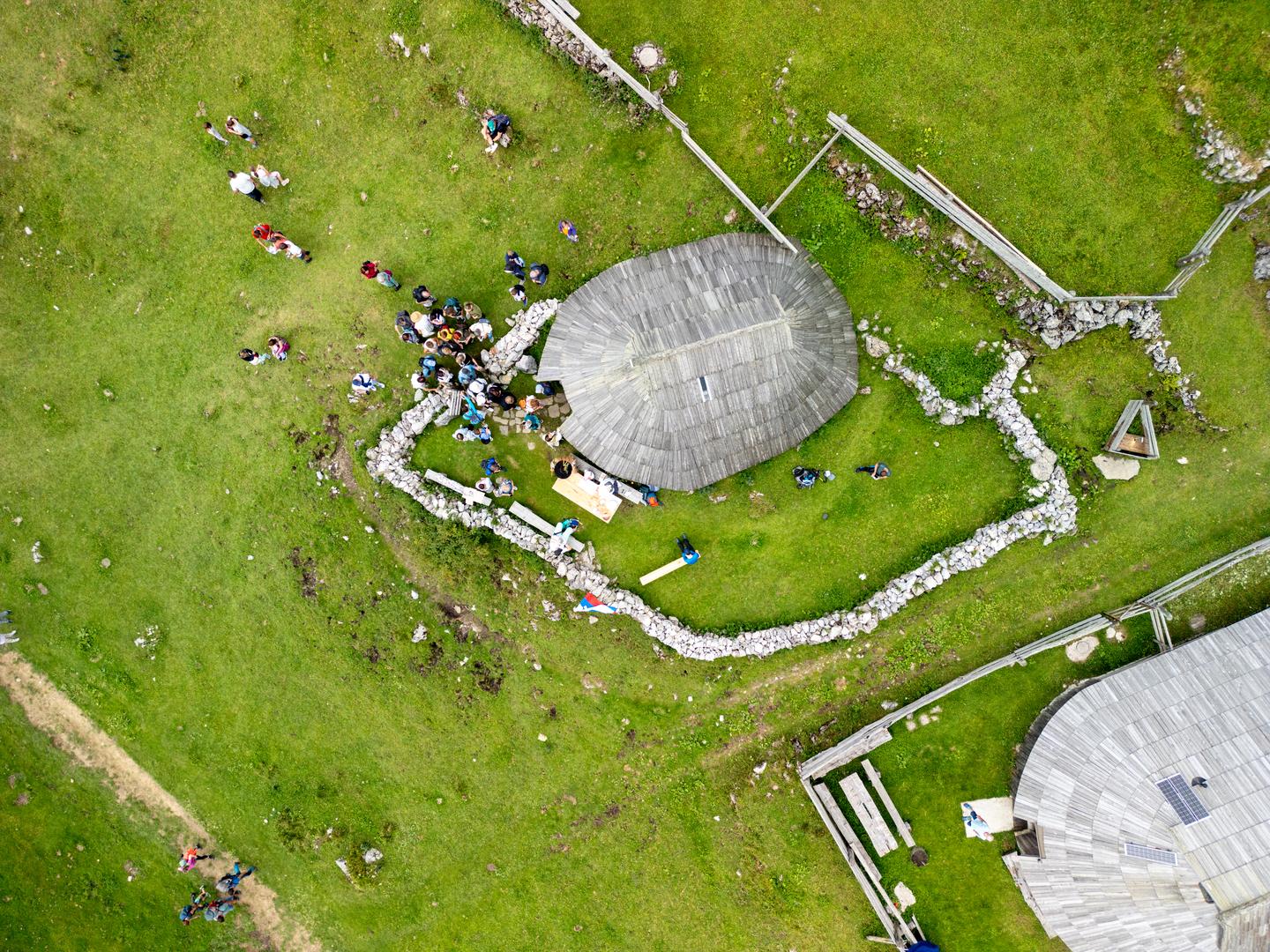Dron posnetek skupine ljudi okoli Preskarjevega muzeja v pastirskem naselju na Veliki planini
